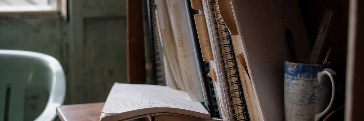 A desk corner and shelf of well-thumbed notebooks and spiral-bound manuals with torn paper page tabs, shot at an angle in window light.